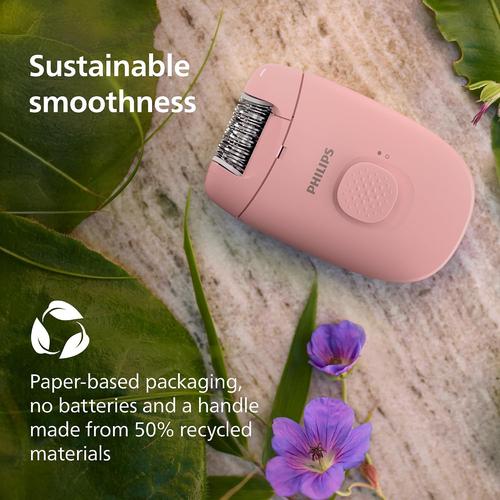 Compact pink Philips corded epilator with a massage cap, displayed on a background of green leaves and purple flowers.