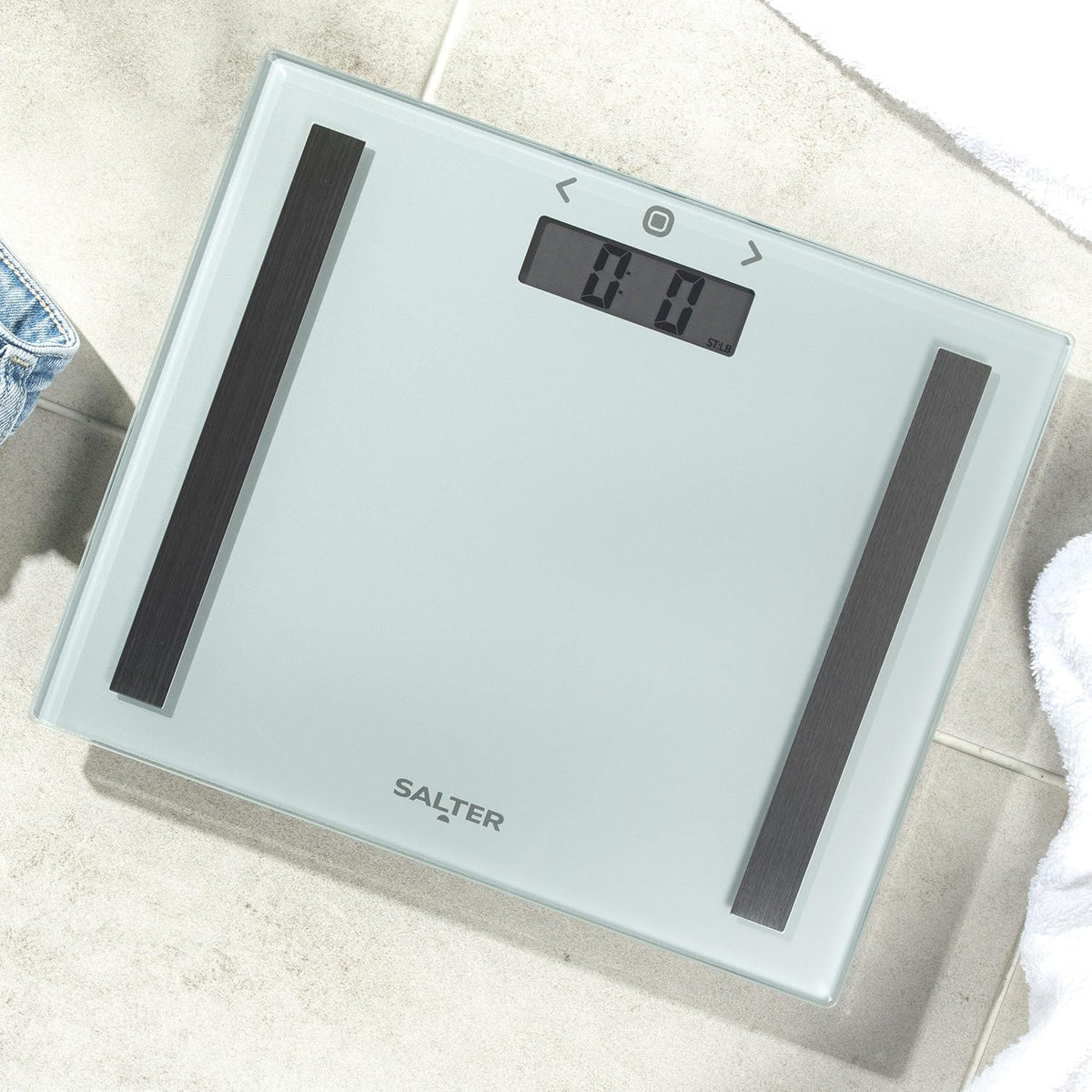 Slim silver Salter digital bathroom scale with glass surface, black sensor strips, and a clear LCD display on tiled floor.